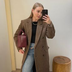 Woman wears brown coat, black top, blue jeans and burgundy bag