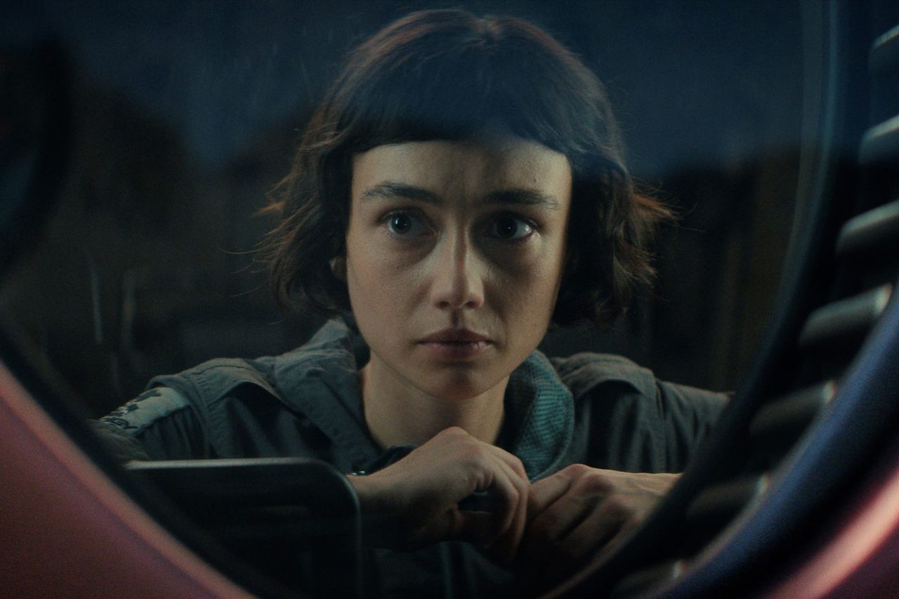 Still from the TV show Alien: Earth. A woman in a gray jumpsuit resting against a window, looking outside.