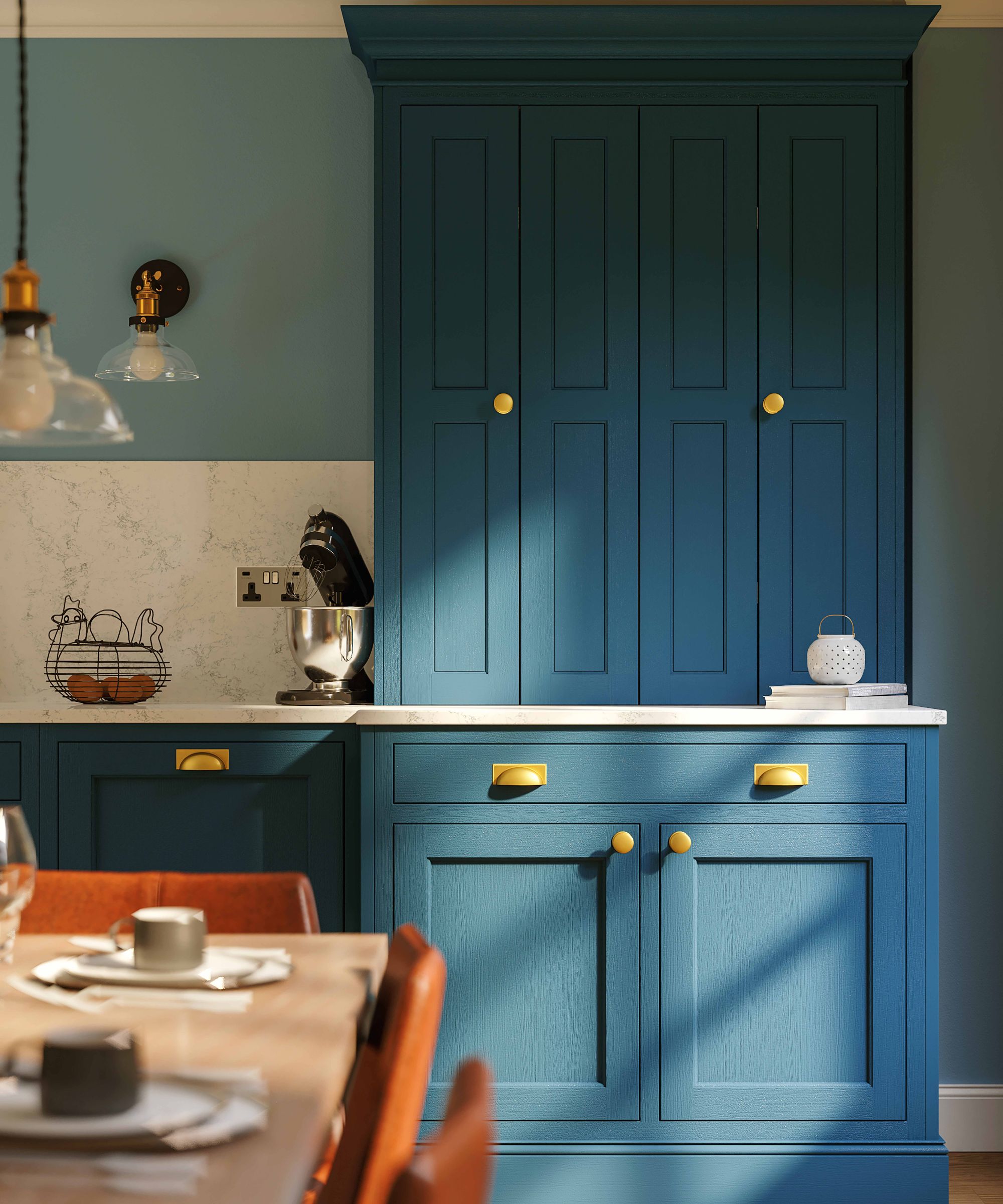 Teal blue in-frame kitchen cabinetry with brass handles, set against soft green walls. A marble worktop runs beneath tall cupboards. Warm sunlight falls across a wooden dining table and leather chairs, creating a calm, elegant, traditional kitchen space.