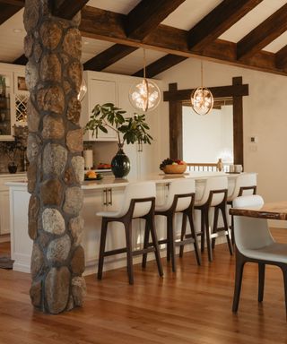 A cream farmhouse dining area with a white breakfast bar with four seats looking onto a cream kitchen, dark wooden beams on the ceiling, and a large stone pillar to the left