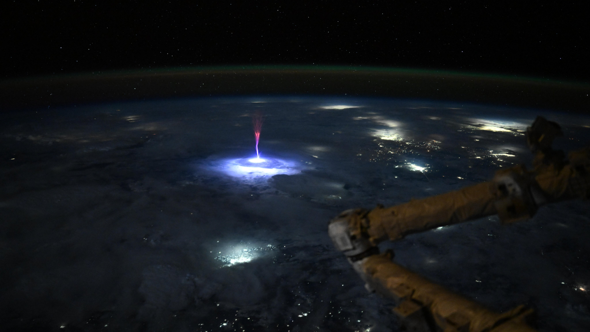A picture of Earth's surface at night captured from the International Space Station. An electrical phenomenon known as a sprite, featuring a tree-like column and red branch-like tendrils can be seen reaching into the upper atmosphere above lightening illuminated clouds.