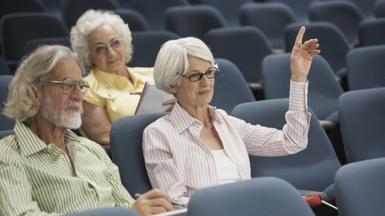 Three older people in an auditorium, and one woman has her hand up to ask a question. 