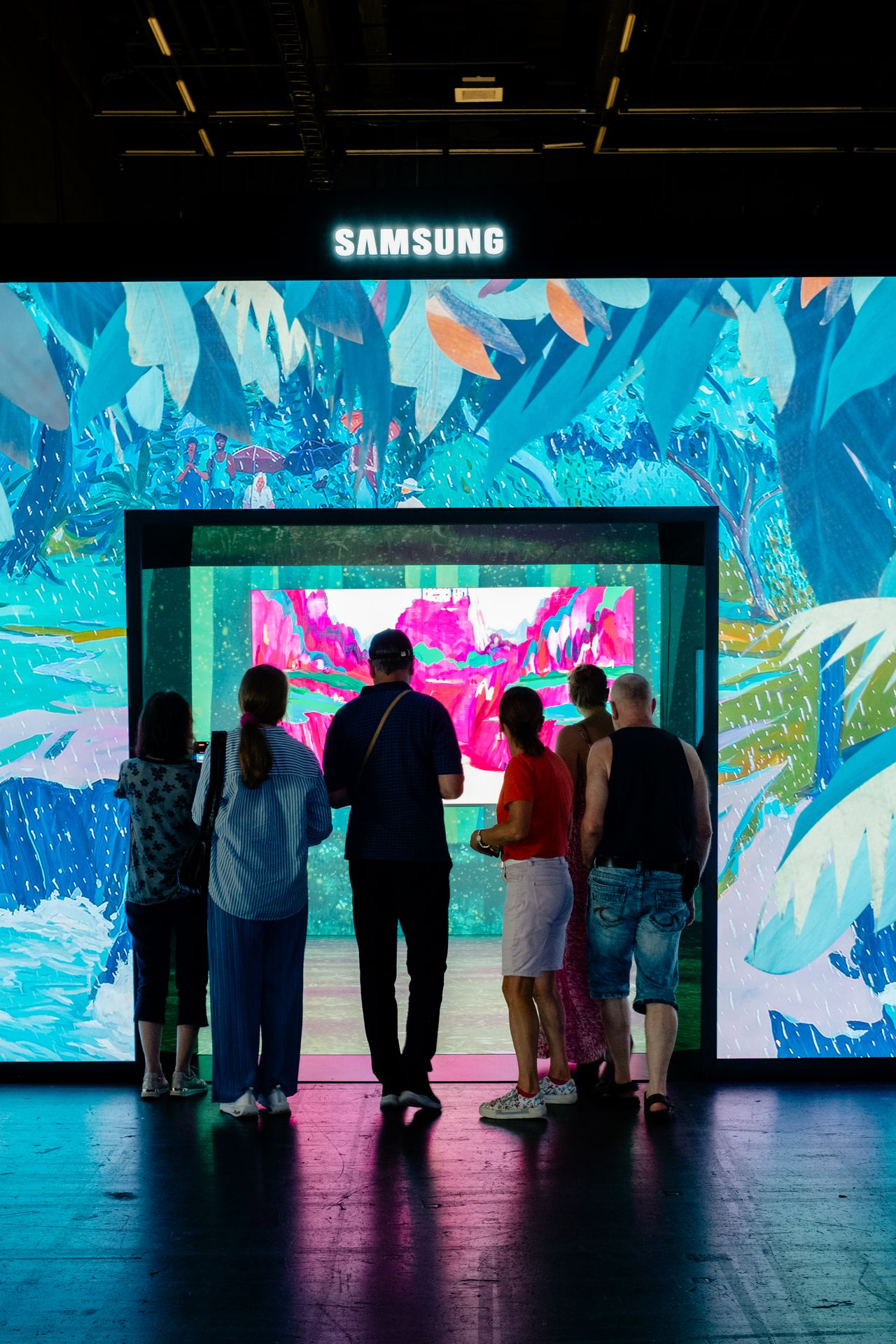 A group of people stands outside an interactive art installation composed of a screens-clad cube projecting paintings.