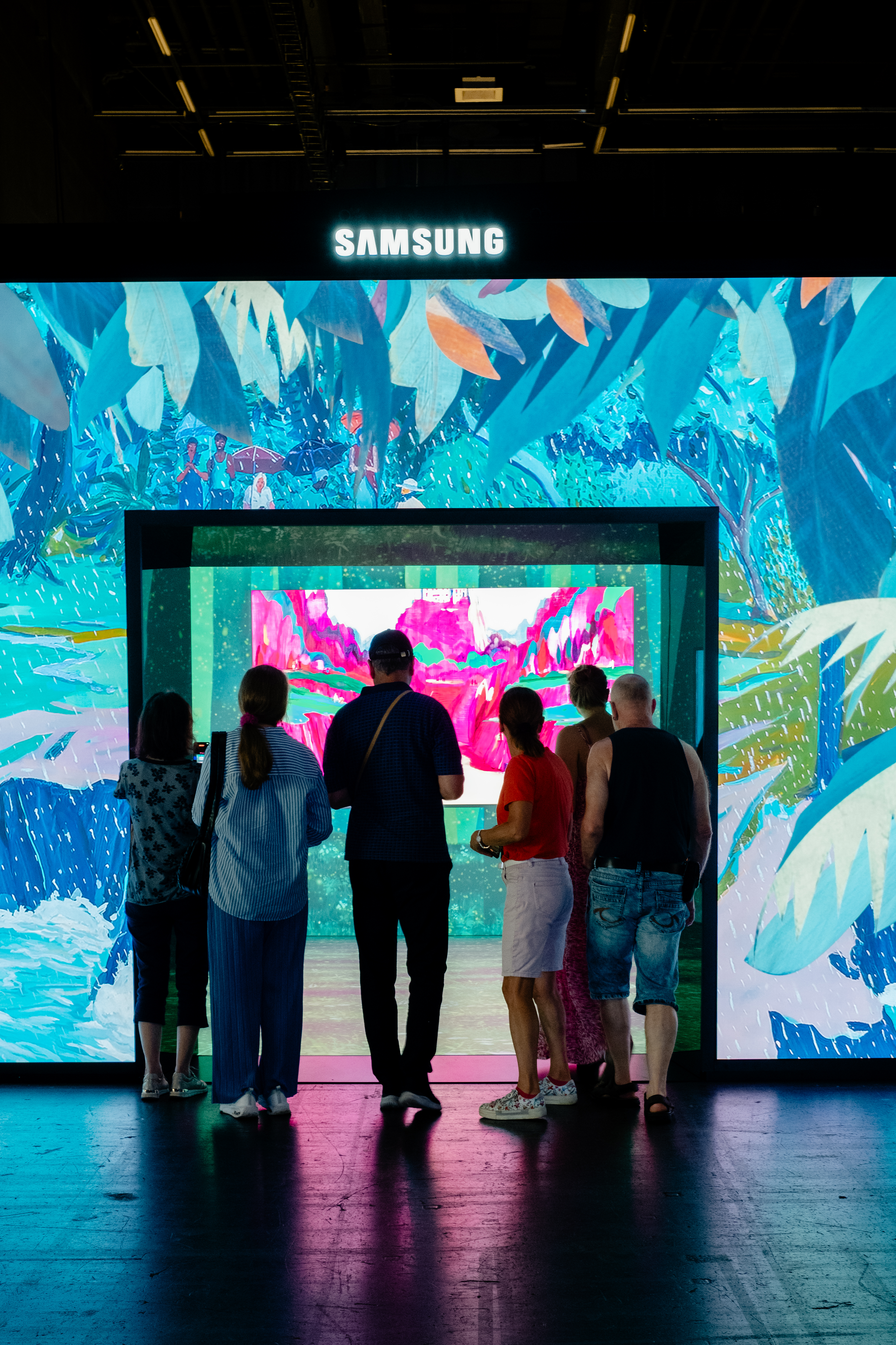 A group of people stands outside an interactive art installation composed of a screens-clad cube projecting paintings.