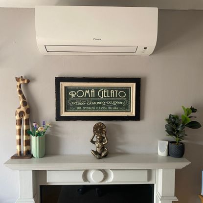 Fitted air conditioning unit above a defunct fireplace with a black and cream picture below, giraffe figure Lego flowers, monkey clock and faux plant on mantlepiece