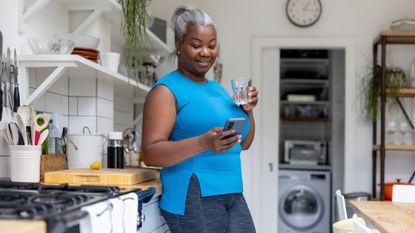 A woman in exercise clothing leans on a kitchen counter looking at her mobile phone. She holds a glass of water.