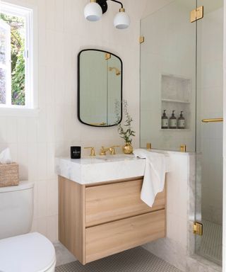 A bathroom with cream colored walls with a window and curved black mirror, a marble sink with a gold tap and wooden base, and a toilet to the left and glass door shower to the right