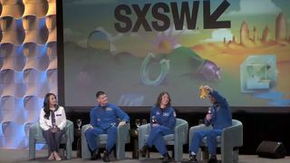 a nasa spokesperson and three astronauts in blue flightsuits sit on a stage at a festival. One of the astronauts holds up a stuffed toy giraffe.