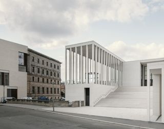 Street level view of the main entrance. The building all white with a grand staircase in the centre and a left section with tall slender pillars.