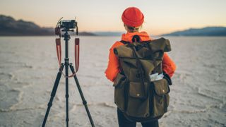 Female photographer wearing backpack watching sky with tripod and camera next to her