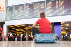 Person sitting on their suitcase, waiting at an arrivals board 