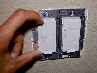 Lutron Caseta in-wall dimmers are shown installed in a wall. A hand rests against one of the devices, positioning it.