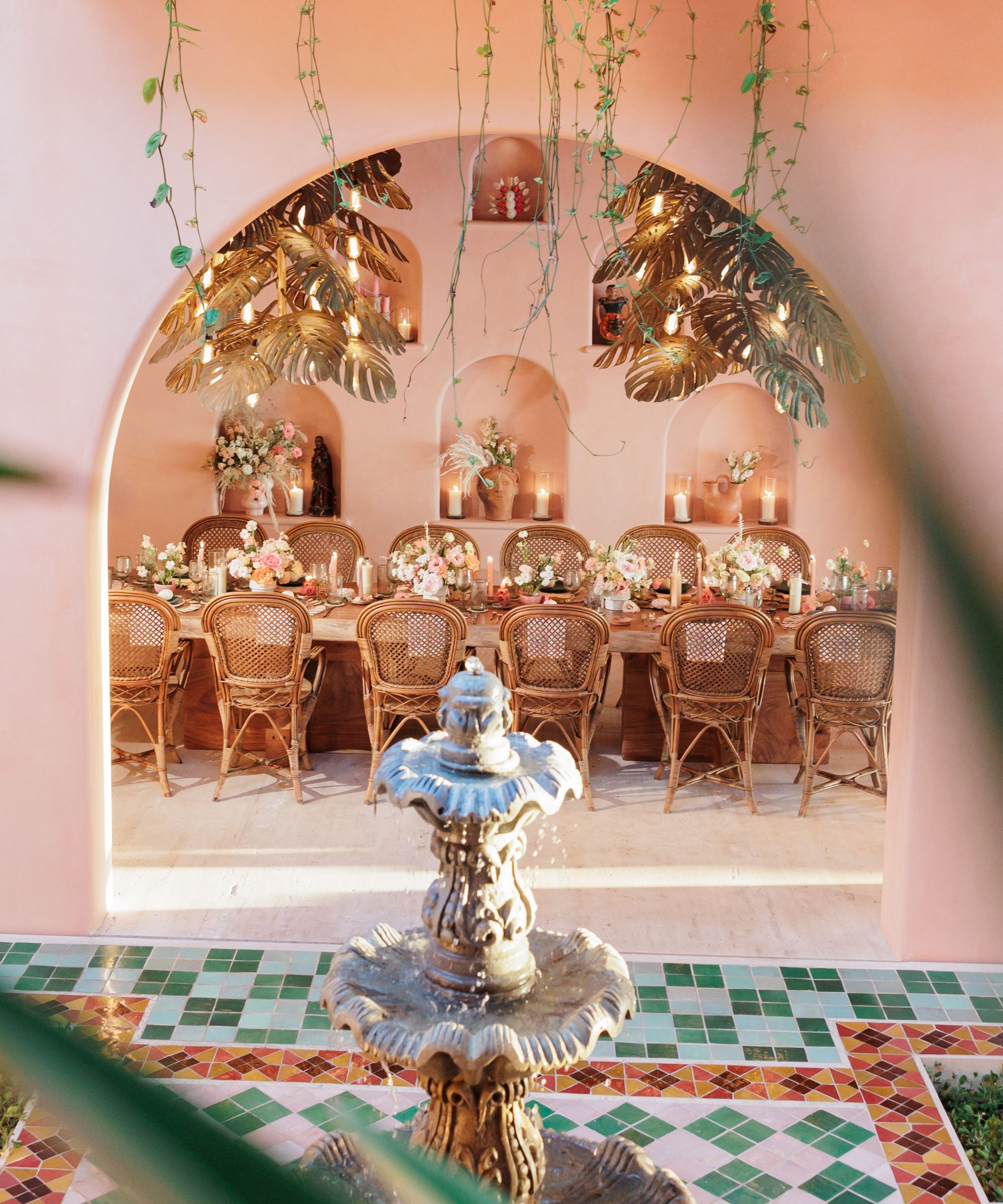 Courtyard dining set up with colorful floor tiles, pink walls and a stone fountain in the foreground and a table within an arch decorated with flowers in the background