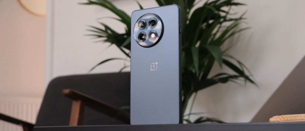 OnePlus 13R phone balanced on a table in a living room with a chair and plant in the background