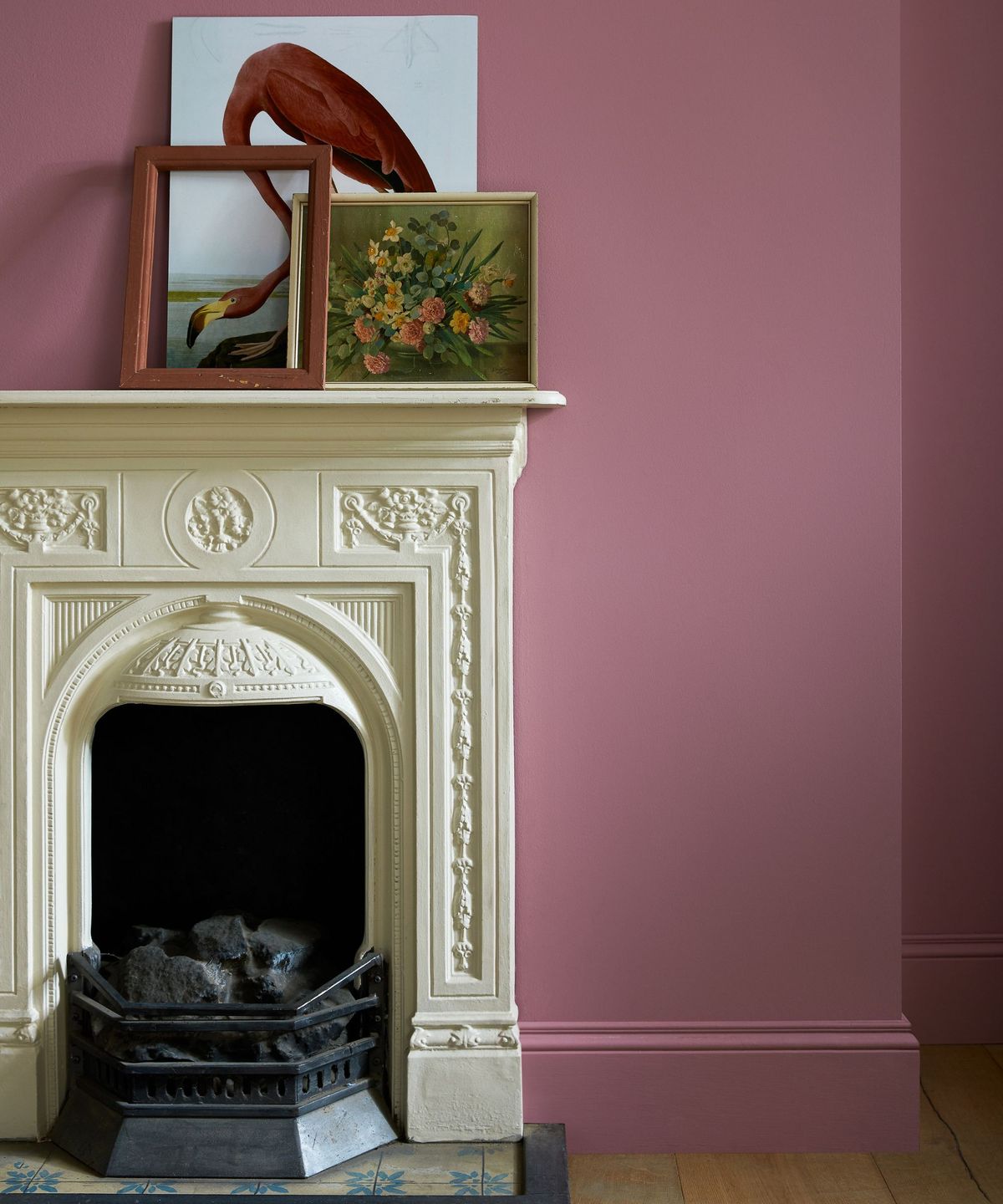 A traditional-style room with a cream ornate fireplace framed by dusky pink walls and matching skirting. Two framed artworks &mdash; one of a flamingo and one floral &mdash; rest casually on the mantelpiece.