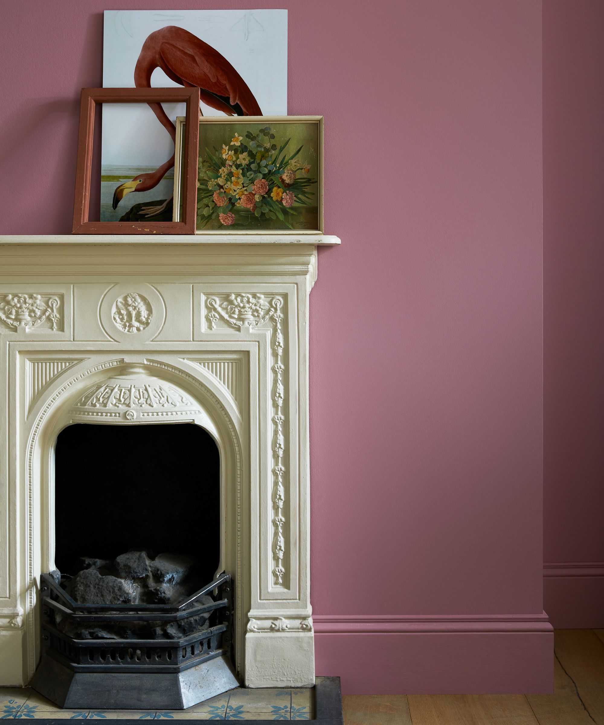 A traditional-style room with a cream ornate fireplace framed by dusky pink walls and matching skirting. Two framed artworks — one of a flamingo and one floral — rest casually on the mantelpiece.