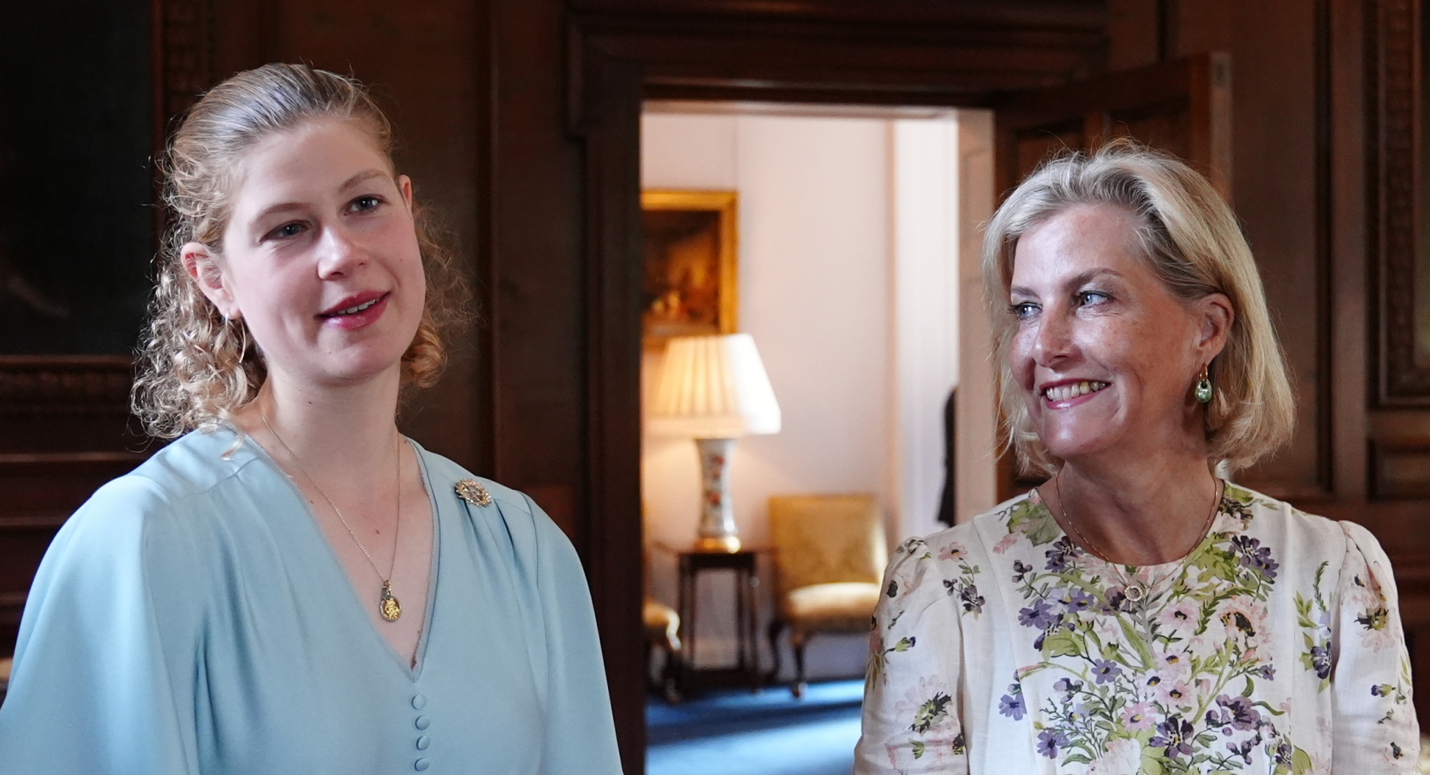 Lady Louise has blonde curly hair and is wearing a light blue dress while her mom Duchess Sophie has a blonde bob and is wearing a floral dress