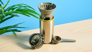 Wacaco Cuppamoka, a stainless steel coffee pour-over brewer, photographed against a blue background, with brown filters and a sippy lid