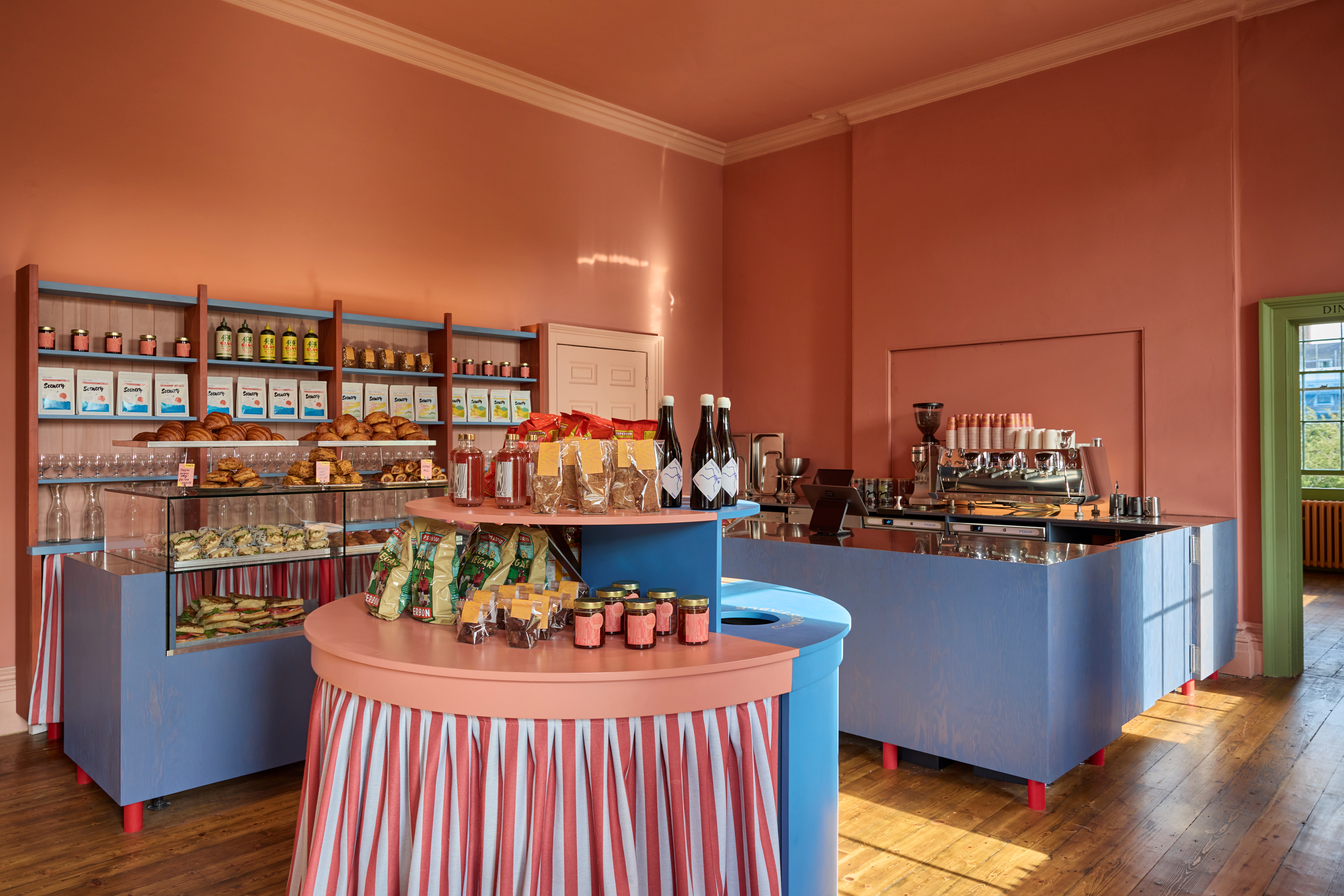 A salmon pink, baby blue, and pea green-tinted deli and cafe set inside a historical building with light wooden floors, decorated with shelves stacked with jars, pastries, and glasses and brought to life by natural light.