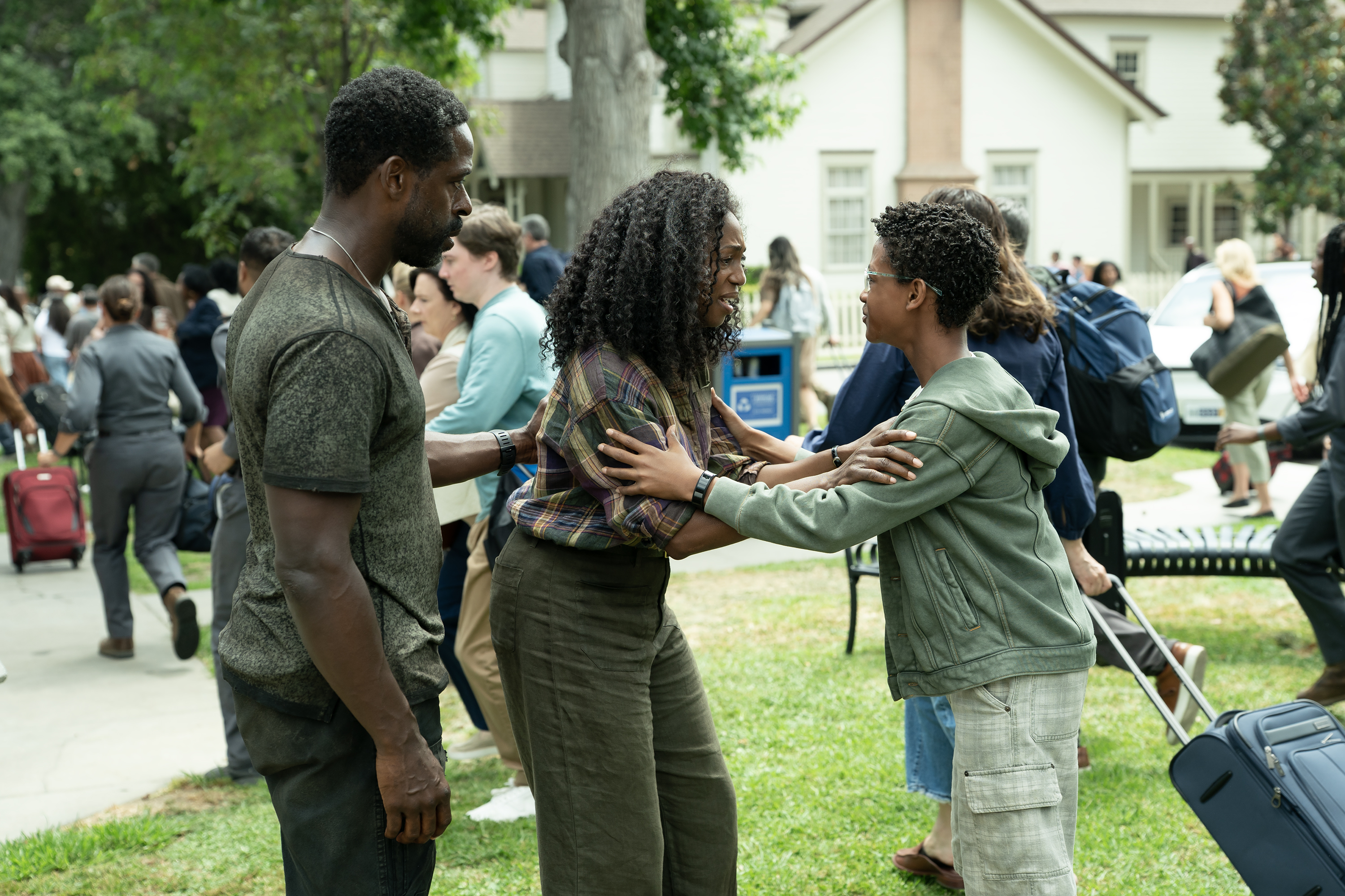 A family (Sterling K. Brown as Xavier Collins, Enuka Okuma as Teri Collins, and Percy Daggs IV as James Collins) holds each other as they stand in front of a crowd of people running with suitcases through a town square, in a still from 'Paradise' season 2.