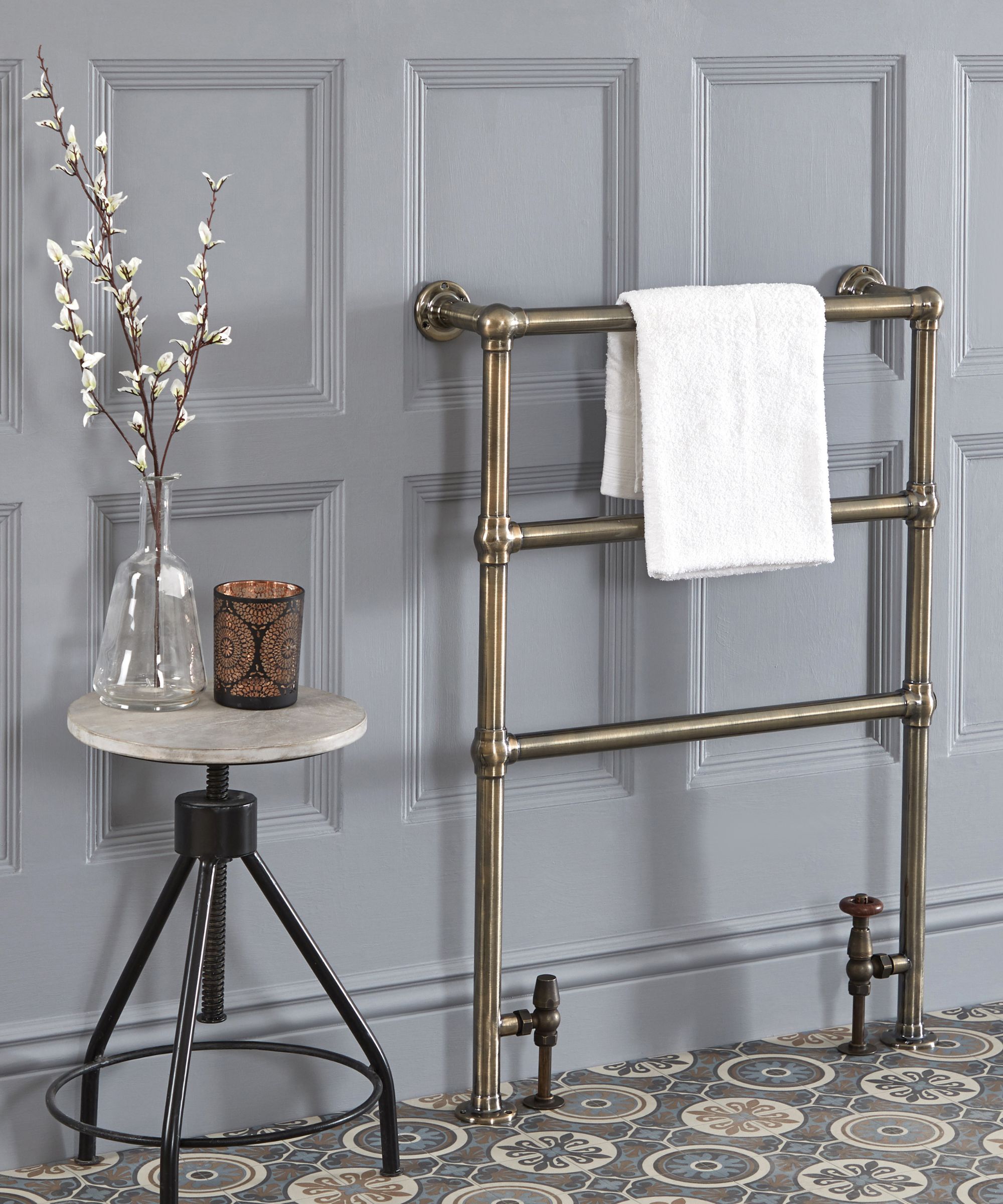 A vintage-style brass towel rail is mounted against grey panelling, styled with a single white towel. Patterned floor tiles and a small side table with glass vase and candle complete the look.