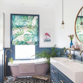 White painted bathroom with blue panelling on the bottom of the wall, a pink freestanding bath, and a white sink unit with blue cabinet underneath