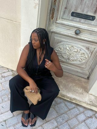 Imani Randolph wearing a black matching vest and trouser set, silver jewelry, black leather flip-flop sandals, and holding a raffia clutch.