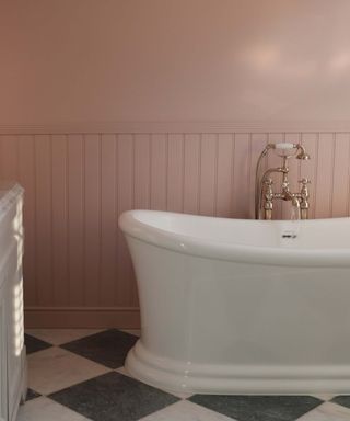 A close-up shot of a bathroom, showing a large freestanding bath standing on a black and white chequered floor. Behind the bath is panelling and a pink painted wall.