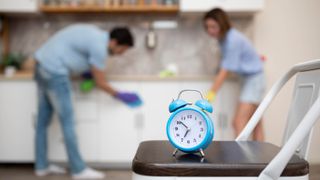 An alarm clock showing the time on a chair with two people cleaning a kitchen in the background
