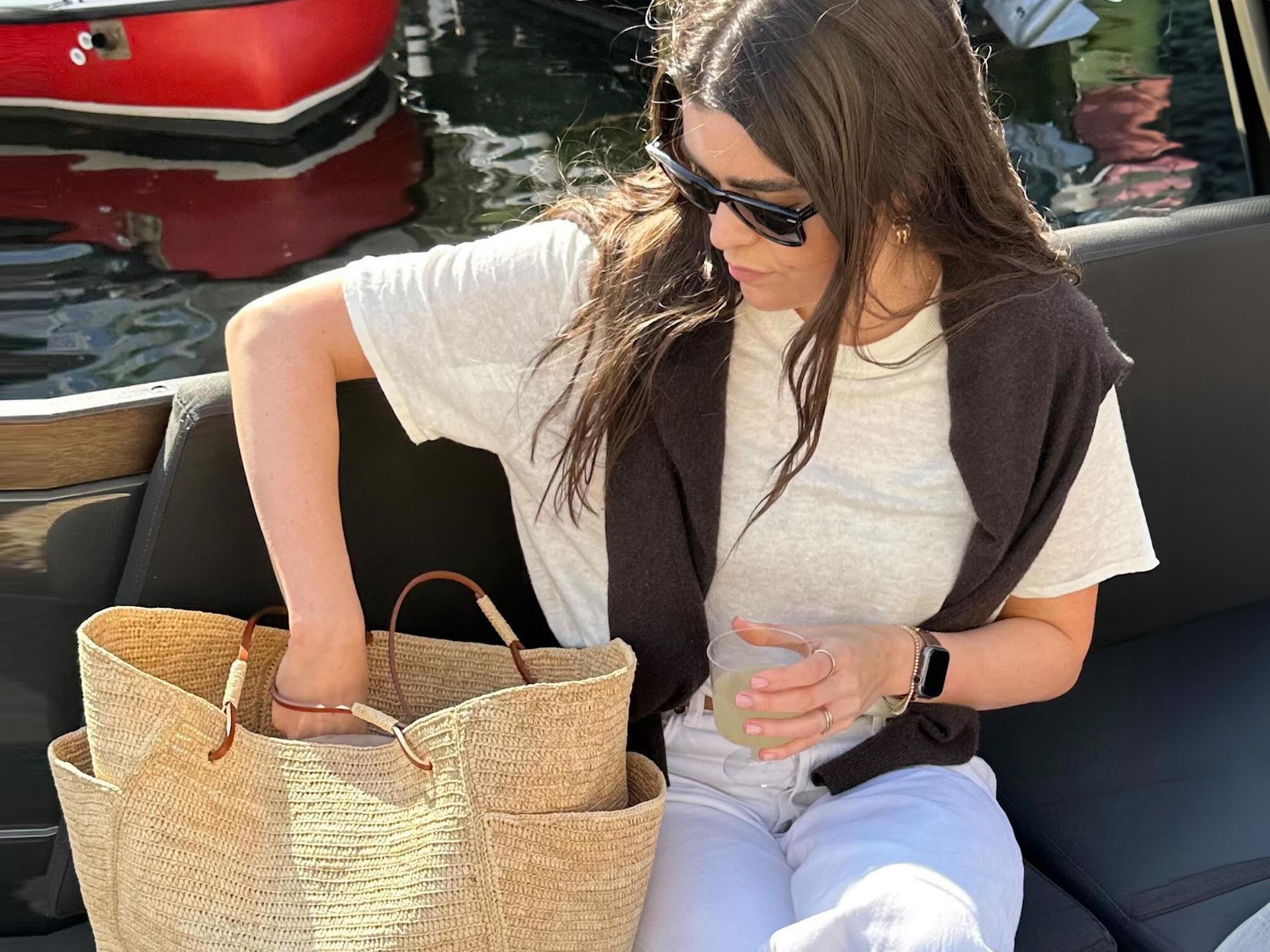 British Anna Newton sitting on a boat with a drink in one hand while looking through her straw tote bag wearing black sunglasses, a linen ivory t-shirt, brown sweater over her shoulders, and white jeans