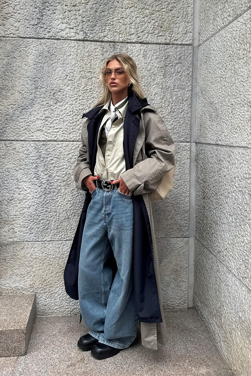 Layering staples for spring 2026 is shown in a street-style photo of a woman standing outside, wearing a tan trench coat over a navy trench coat. The rest of the look includes three contrasting layered button-down shirts tucked into baggy jeans styled with a black Gucci belt, black ankle boots, and aviator sunglasses
