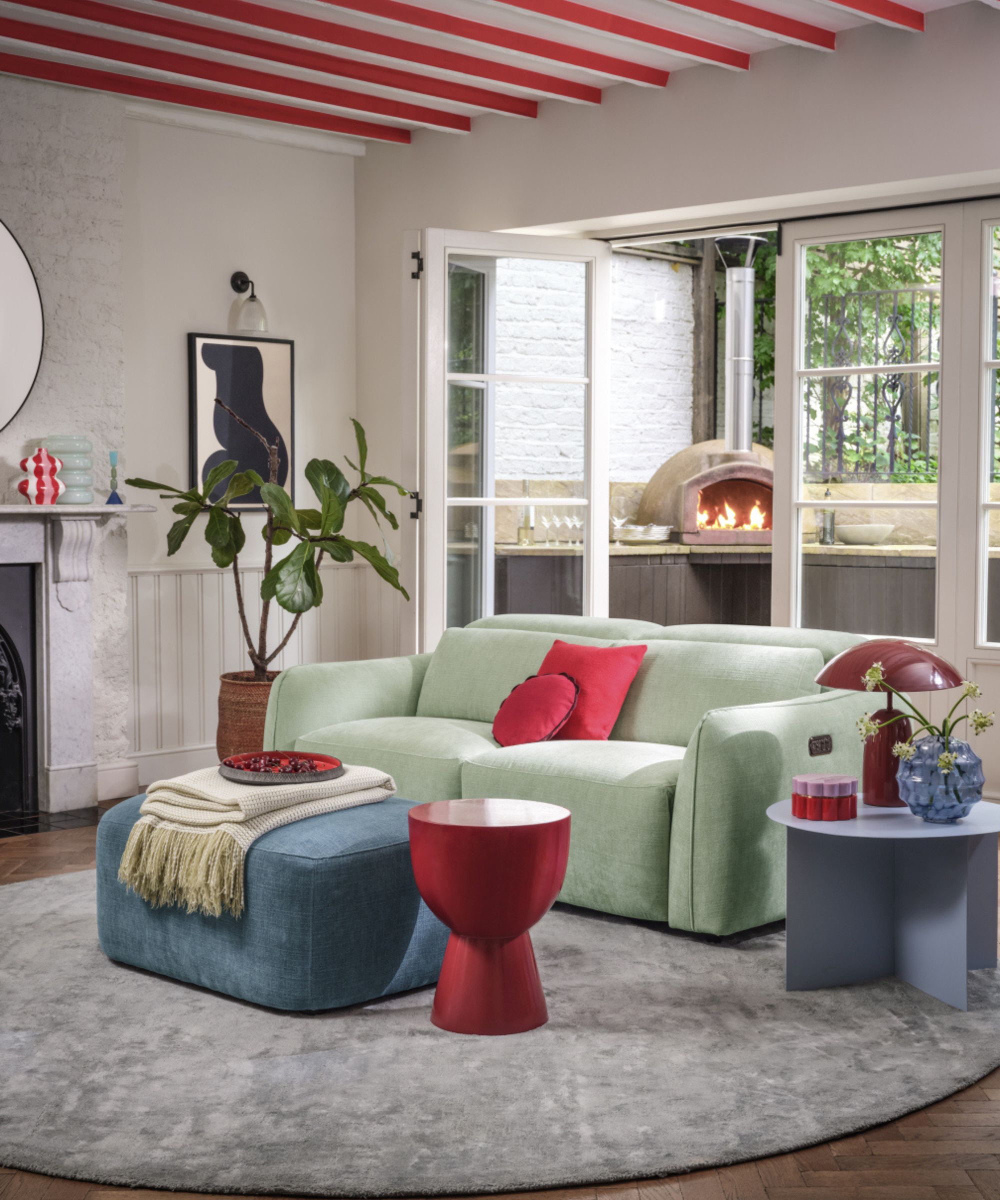 Small cottage style living room with red painted ceiling beams and matching accents including cushions, side tables, conical lamp and mantel decor. The French doors are open into a small courtyard with pizza oven blazing