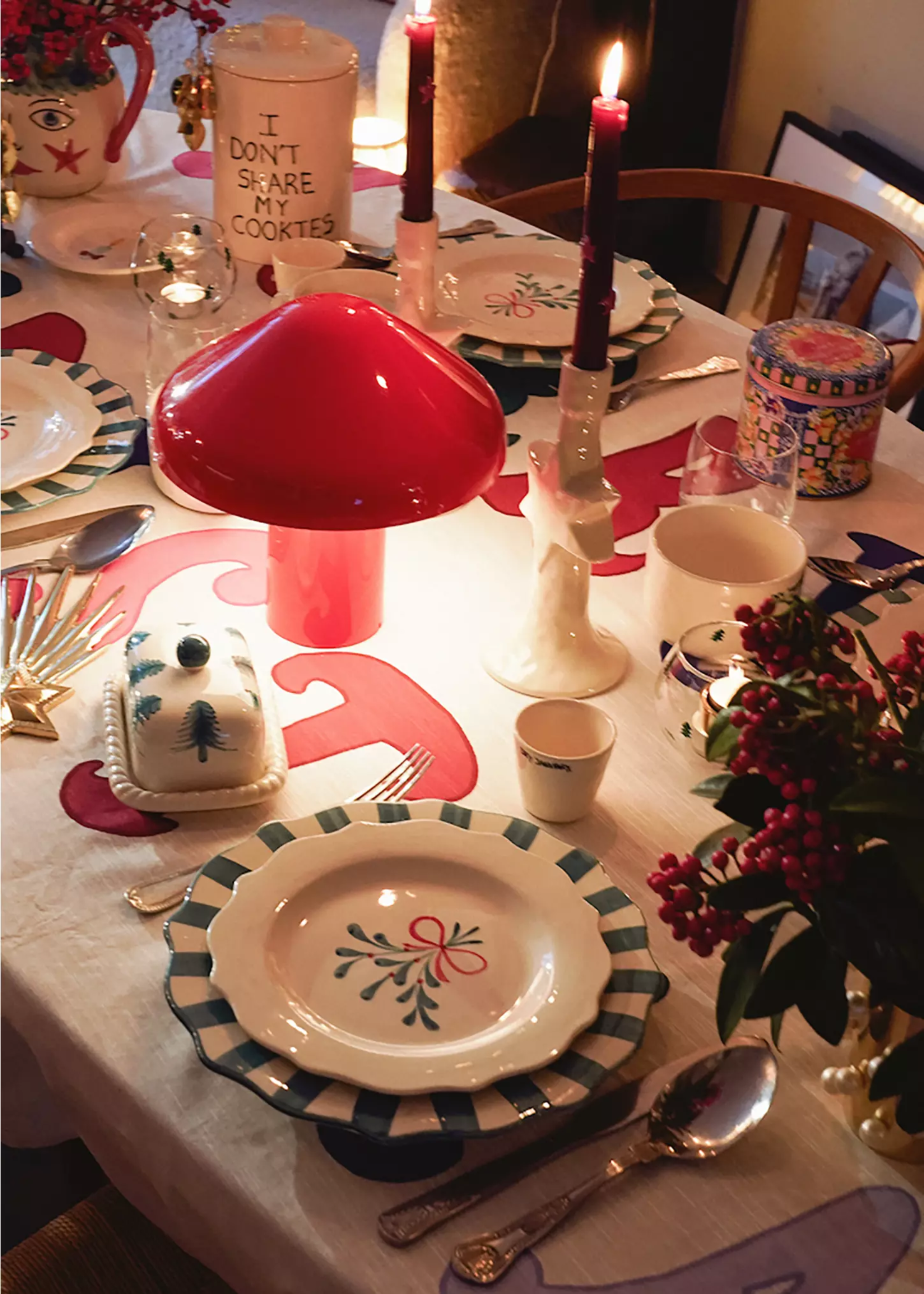 A table set for Christmas with a red table lamp, layered plates, lit candles in tapers and a festive floral arrangement
