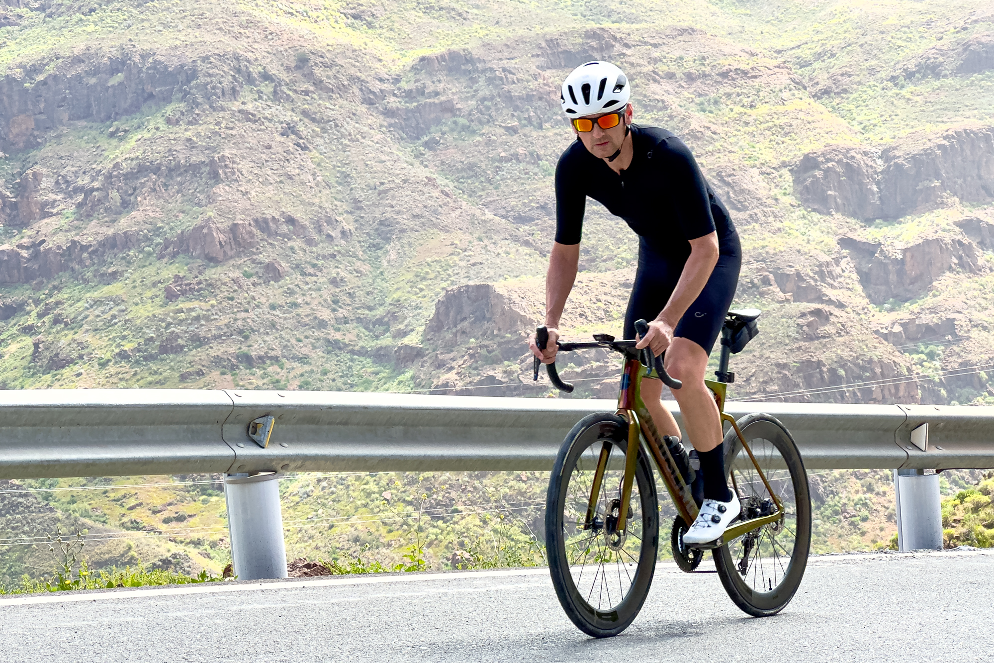 man wearing a black jersey, sunglasses and a white helmet standing up riding a bike