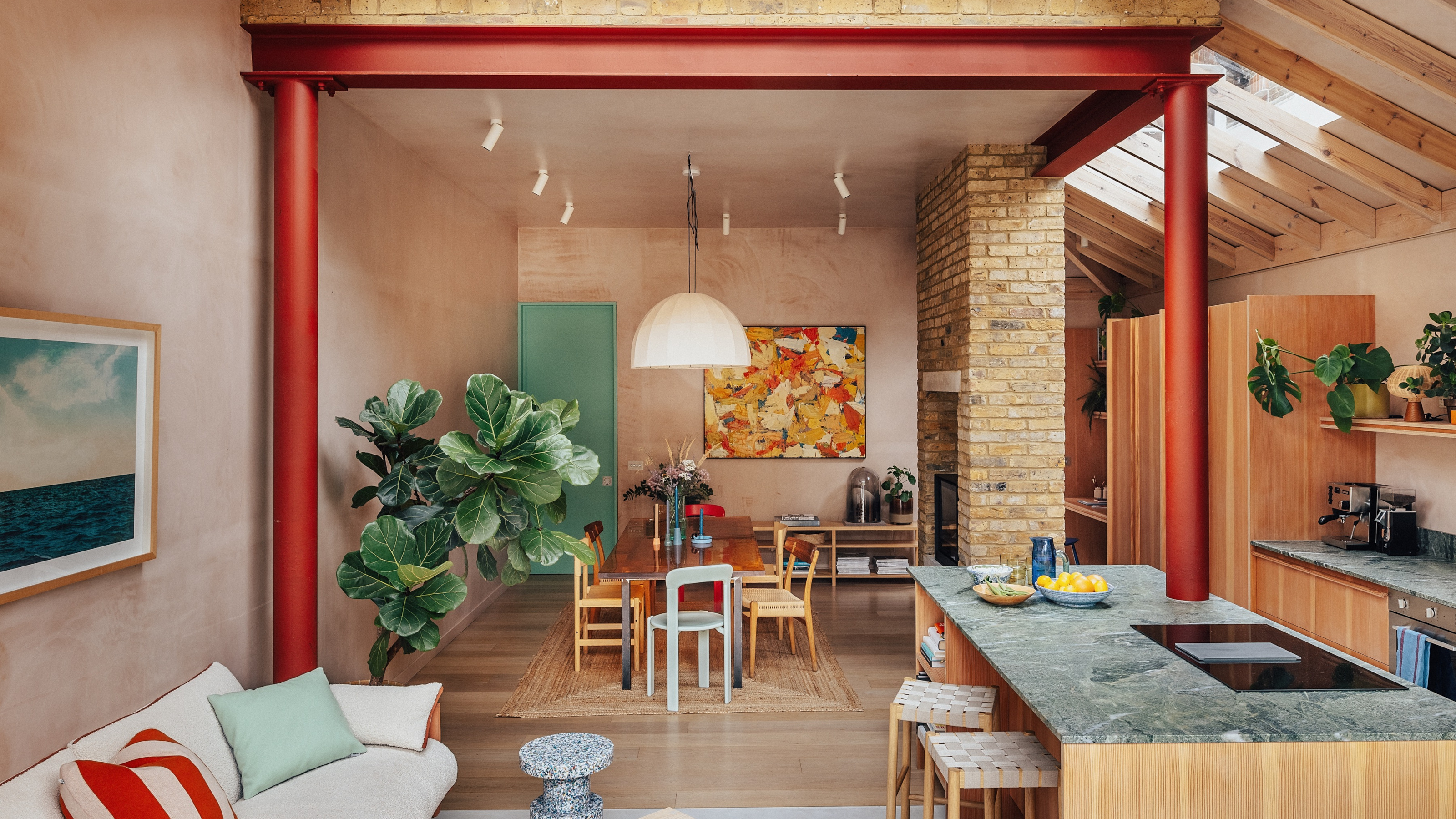 Open-plan kitchen-diner with plaster pink walls, wood floor, wood kitchen cabinetry with green stone worktop, red structural steel, exposed brick fireplace