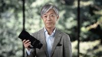 Sigma CEO Kazuto Yamaki, wearing a grey suit, holding up a lens, against a defocused nature background