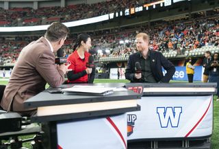 Prince Harry sitting at a sports commentator desk holding a mic with a man and woman