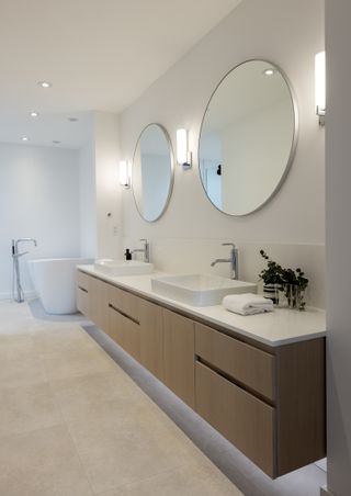 A modern bathroom with a double vanity with wooden cabinetry and white counter top, with two circular mirrors above