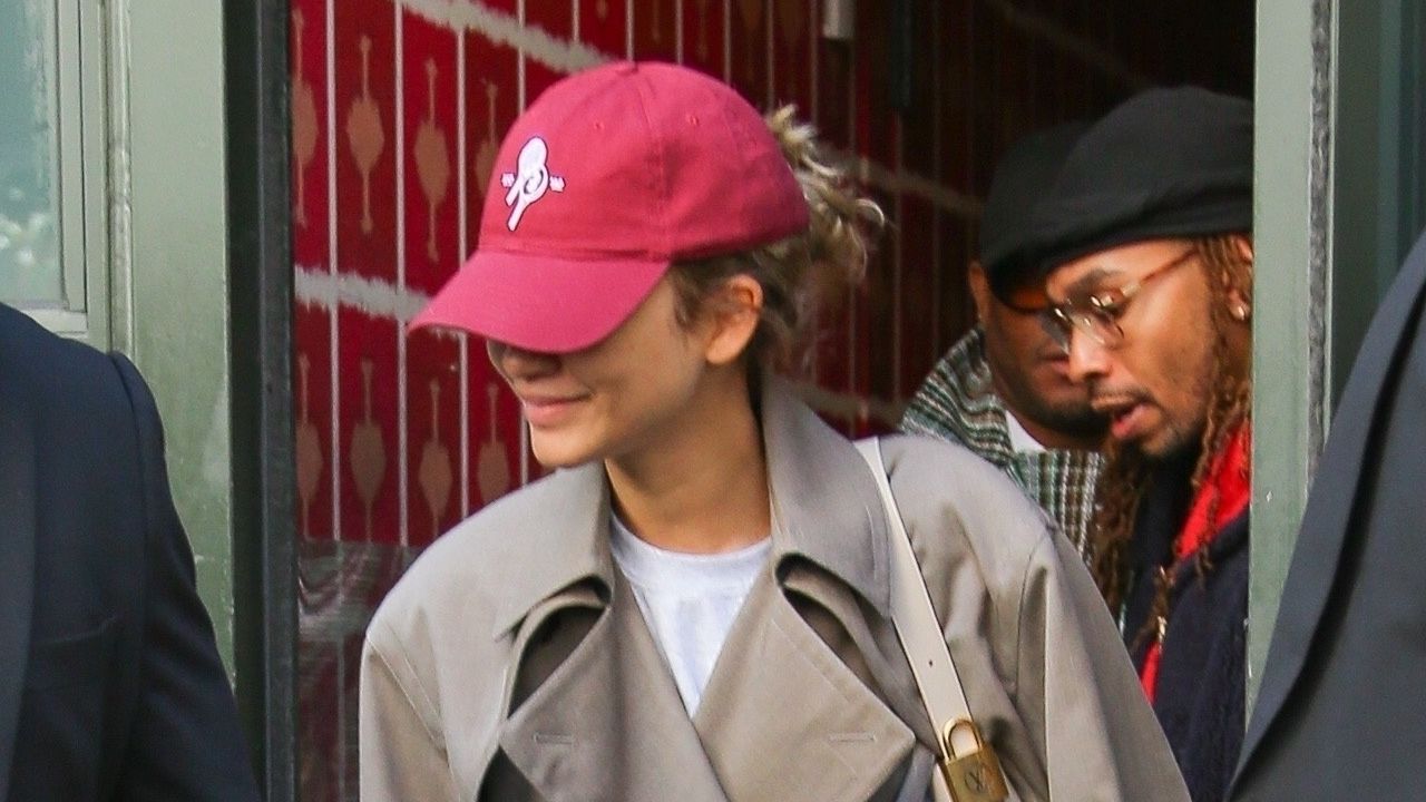 Zendaya wearing a trench coat, white tee, black pants, sneakers, and a red baseball cap while out in New York City April 2024