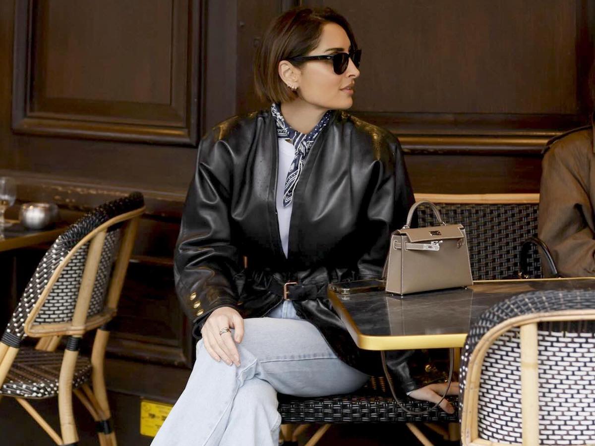 a stylish French woman sits at an outdoor cafe table with her legs crossed wearing a black leather jacket, tied neck scarf, mini Hermes bag, and jeans