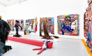 Art gallery with white walls, ceiling and grey flooring featuring colourful wall canvases and grotesque sculptures and knitted patchwork dolls
