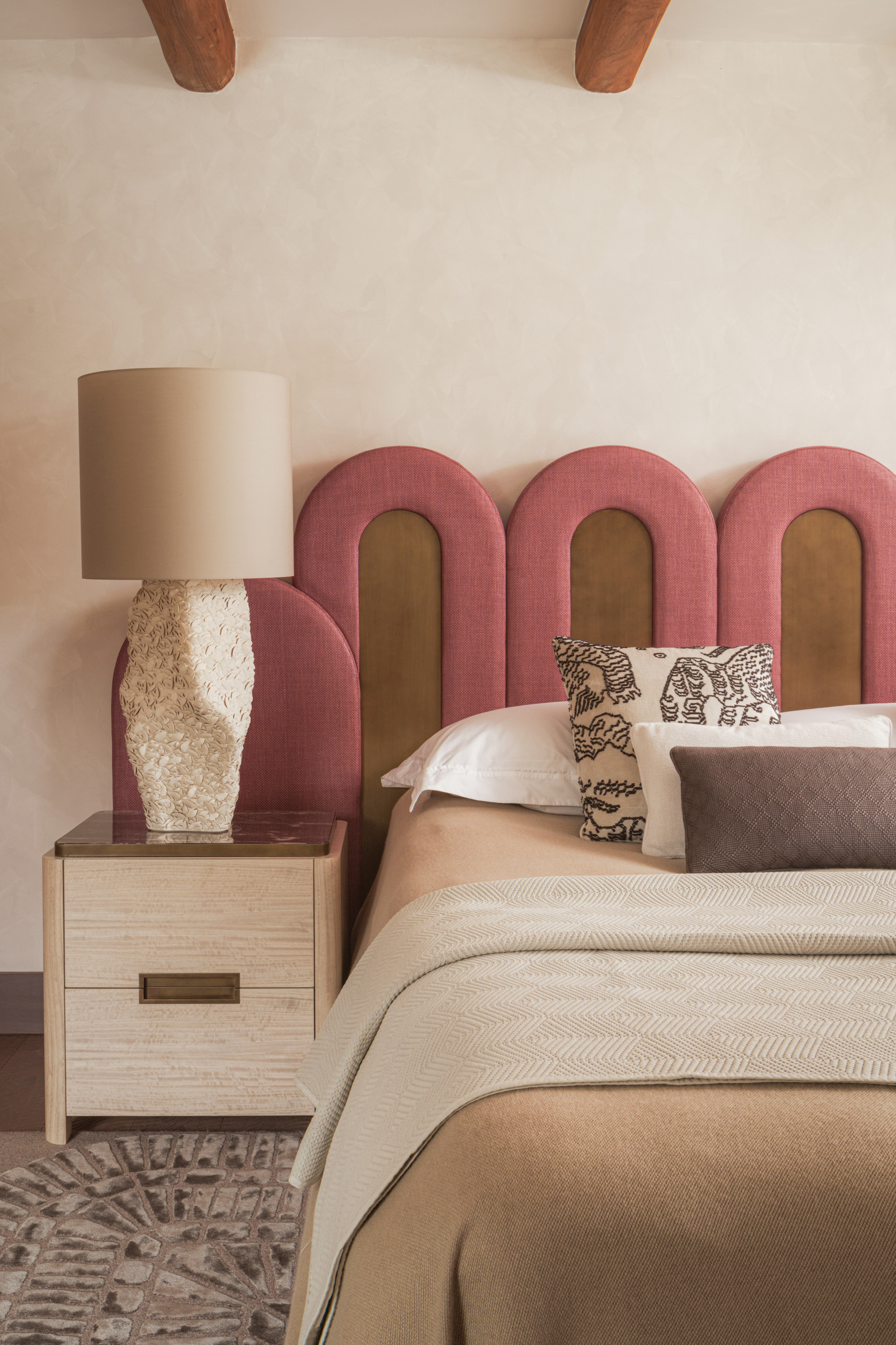Bedroom with pink and brown curved headboard, beige bedside table and lamp, brown bed linen