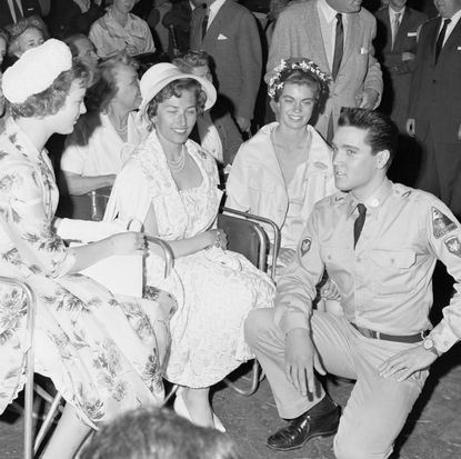 A black-and-white photo of Elvis Presley wearing a military uniform kneeling in front of Princesses Margrethe of Denmark, Astrid of Norway, and Margaretha of Sweden.