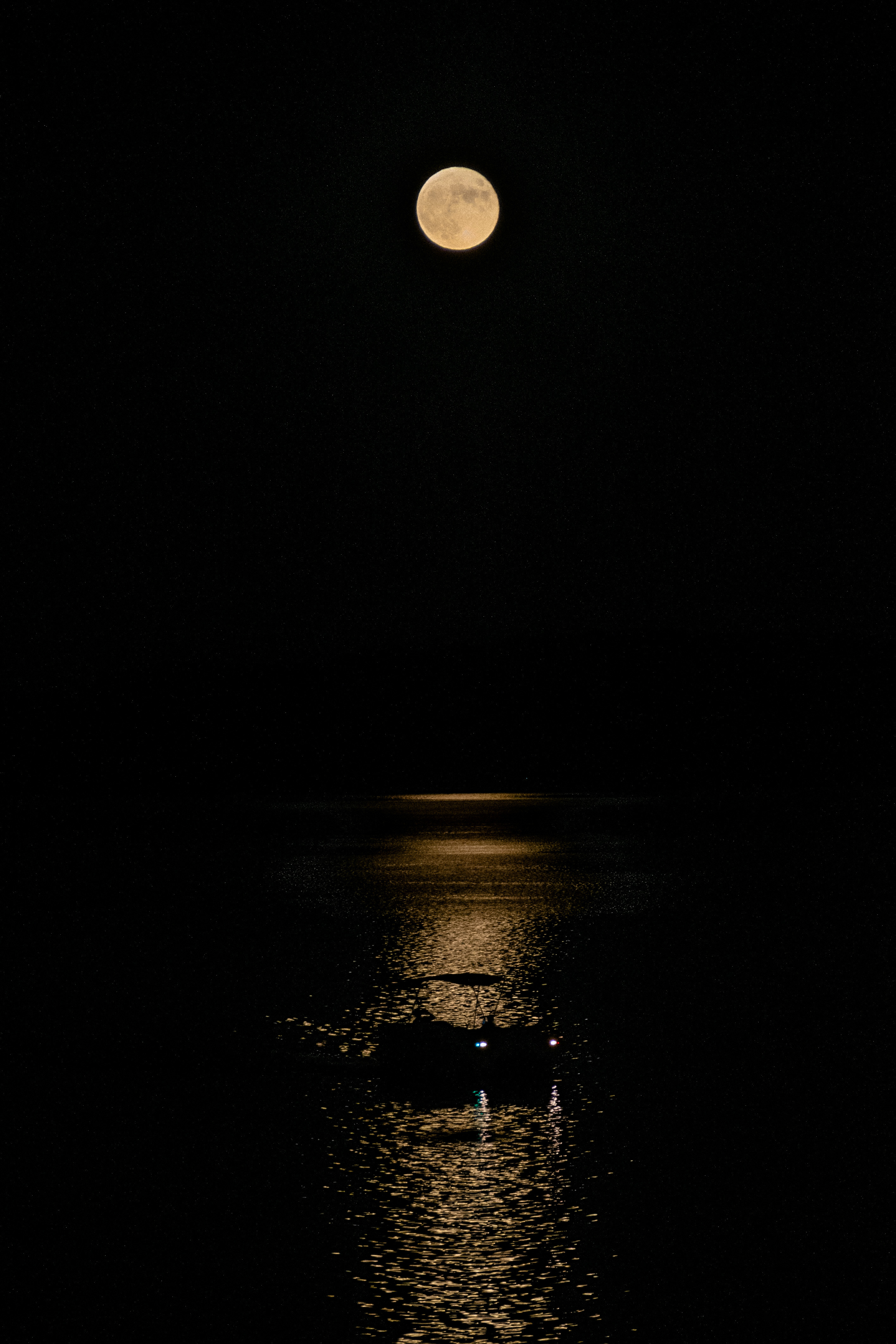A full moon rises against the black sky, and a boat passes by its long, narrow reflection on the water.