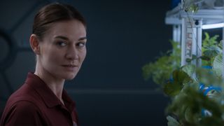 A woman is glancing to a person off screen. She is standing in front of an indoor garden shelf.