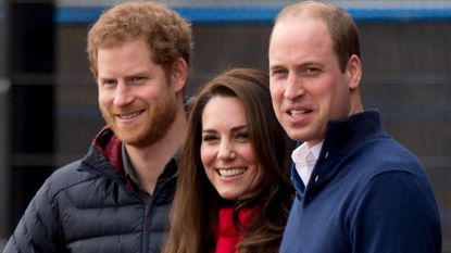 Prince Harry, Princess Kate and Prince William wearing coats standing next to each other and smiling at the camera