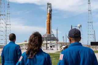 three people in blue flight suits look up at a massive orange rocket on a launch pad