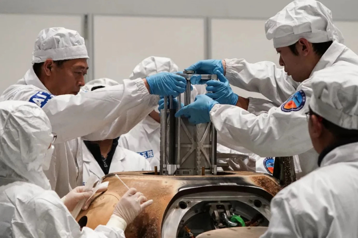 lab technicians in white clean suits lift a metal container out of a charred space capsule