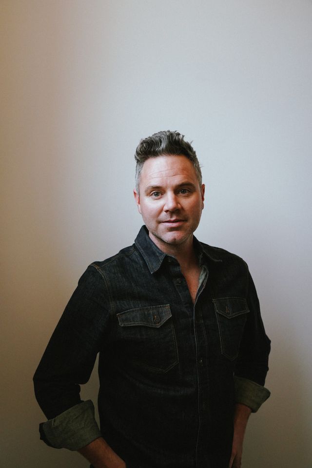 Man standing with black shirt and hands in pockets looking at teh camera.