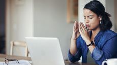 Woman sits in front of laptop at a table in a domestic setting. She has her eyes closed, her lips are pursed as if exhaling and her hands are held close to her face.